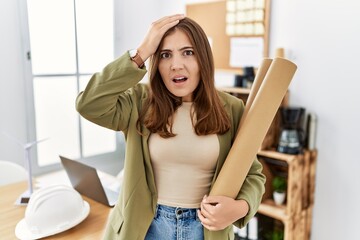 Young brunette woman holding paper blueprints at the office stressed and frustrated with hand on...