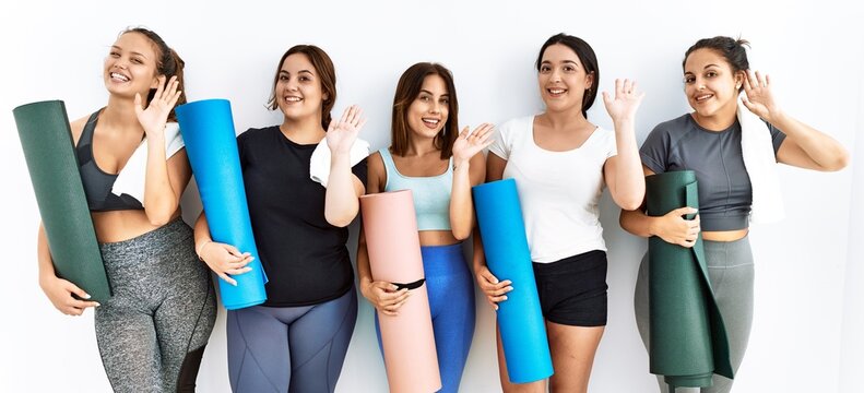 Group of women holding yoga mat standing over isolated background waiving saying hello happy and smiling, friendly welcome gesture