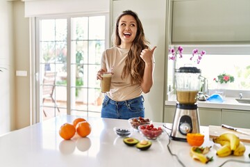 Beautiful young brunette woman drinking glass of smoothie at the kitchen pointing thumb up to the side smiling happy with open mouth