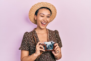 Beautiful hispanic woman with short hair wearing summer hat holding vintage camera winking looking...