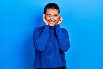 Beautiful hispanic woman with short hair wearing turtleneck sweater covering ears with fingers with annoyed expression for the noise of loud music. deaf concept.