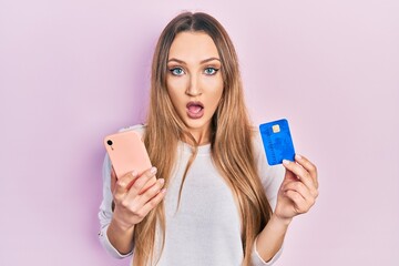 Young blonde girl holding smartphone and credit card afraid and shocked with surprise and amazed expression, fear and excited face.
