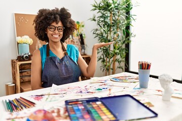Beautiful african american woman with afro hair painting at art studio smiling cheerful presenting and pointing with palm of hand looking at the camera.