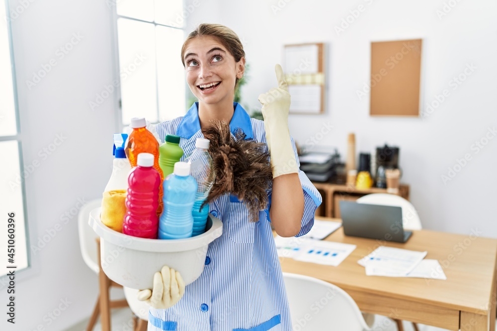 Sticker Young blonde woman wearing cleaner uniform holding cleaning products smiling amazed and surprised and pointing up with fingers and raised arms.