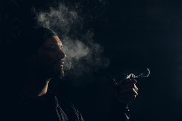 Man smokes smoking pipe against dark background. Back side lit profile portrait with smoke