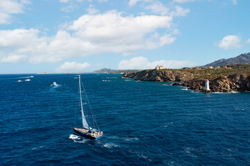 View from above, stunning aerial view of a sailboat sailing on a rough sea in the foreground and a rocky coastline with a lighthouse in the distance. Cala del faro, Sardinia, Italy.