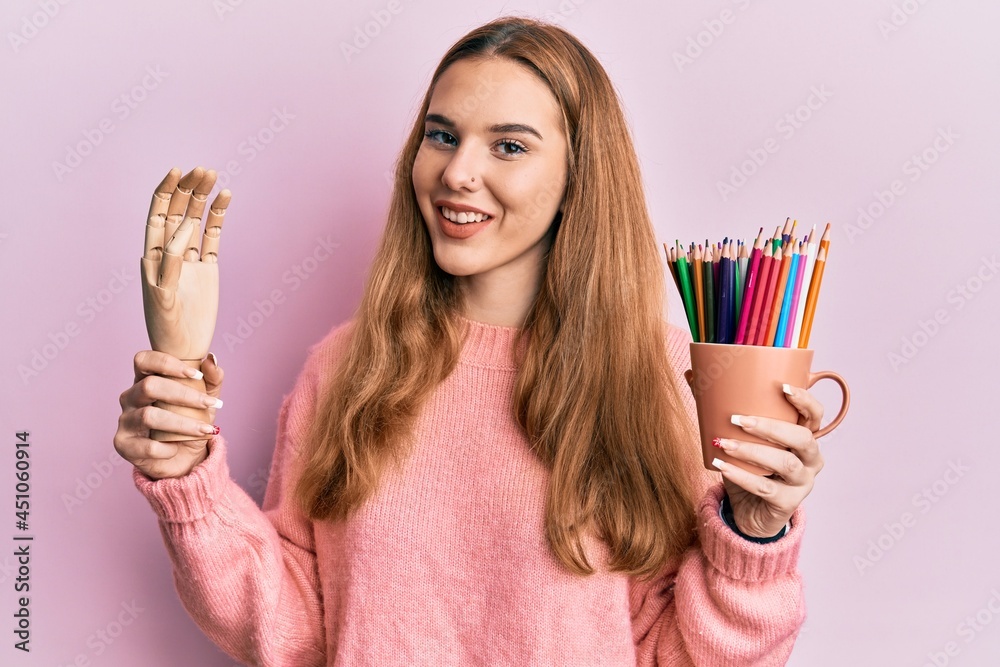 Sticker young blonde woman holding small wooden manikin hand and pencils winking looking at the camera with 