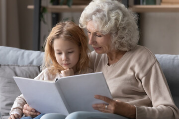 Happy loving Caucasian mature grandmother sit rest on sofa in living room read book with cute little teen granddaughter. Caring grandparent enjoy literature with small grandchild. Hobby concept.