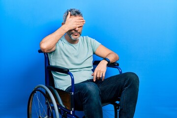 Fototapeta premium Handsome middle age man with grey hair sitting on wheelchair covering eyes with hand, looking serious and sad. sightless, hiding and rejection concept