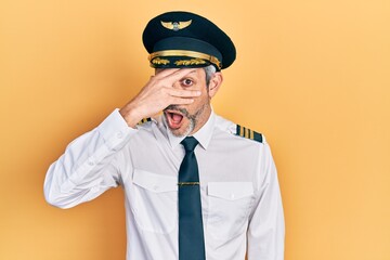 Handsome middle age man with grey hair wearing airplane pilot uniform peeking in shock covering face and eyes with hand, looking through fingers with embarrassed expression.