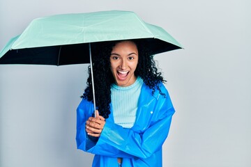 Young hispanic woman with curly hair wearing a raincoat and umbrella smiling and laughing hard out loud because funny crazy joke.