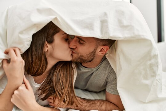 Young caucasian couple covering with bed sheet and kissing on the bed at home.