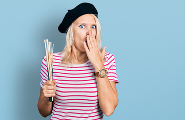 Young caucasian woman holding paintbrushes covering mouth with hand, shocked and afraid for...