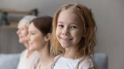 Portrait of smiling small teen girl child offspring stand forefront of young mother and old grandmother. Happy three generations of Caucasian women pose together show family unity and bonding.