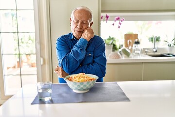 Senior man with grey hair eating pasta spaghetti at home thinking looking tired and bored with...