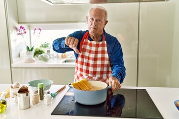 Senior man with grey hair cooking spaghetti at home kitchen with angry face, negative sign showing dislike with thumbs down, rejection concept