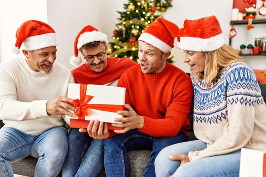 Group of middle age friends holding gifts sitting on the sofa by christmas tree at home.