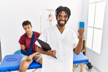 Young hispanic man working at pain recovery clinic with a man with broken arm very happy and excited doing winner gesture with arms raised, smiling and screaming for success. celebration concept.