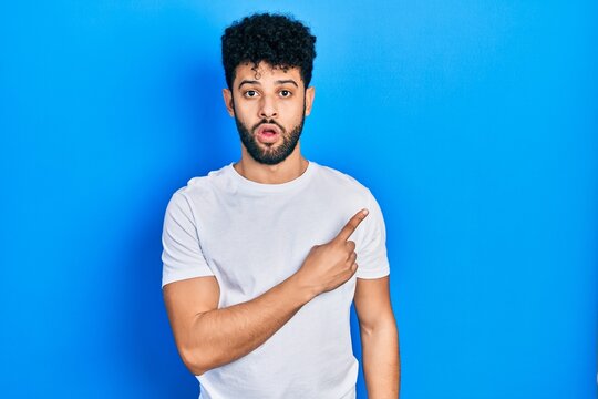 Young arab man with beard wearing casual white t shirt surprised pointing with finger to the side, open mouth amazed expression.