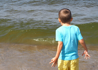 a five-year-old boy in a blue T-shirt and yellow shorts stands on the beach against the background of the sea . rear view. seashore