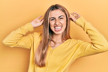 Beautiful hispanic woman wearing casual yellow sweater smiling pulling ears with fingers, funny...
