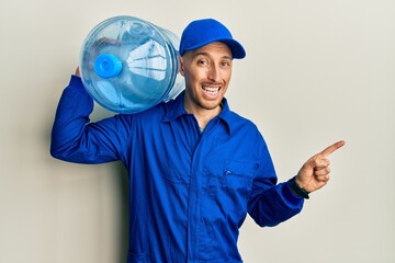 Bald courier man with beard holding a gallon bottle of water for delivery smiling happy pointing with hand and finger to the side