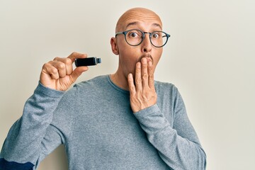 Bald man with beard holding removable memory usb covering mouth with hand, shocked and afraid for...