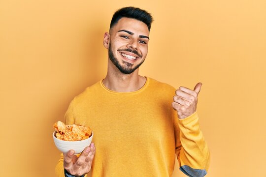 Young hispanic man with beard holding nachos potato chips pointing thumb up to the side smiling happy with open mouth