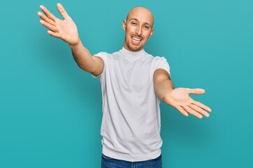 Bald man with beard wearing casual white t shirt looking at the camera smiling with open arms for hug. cheerful expression embracing happiness.
