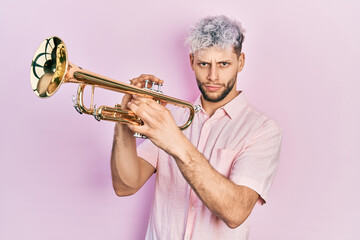 Young hispanic man with modern dyed hair playing trumpet skeptic and nervous, frowning upset...