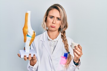 Beautiful young blonde doctor woman holding anatomical model of knee joint and protein powder...