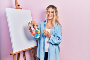 Beautiful young blonde woman standing drawing with palette by painter easel stand winking looking...