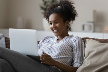 Smiling African American woman relax on sofa talk speak on video call on laptop. Happy young ethnic female have fun laugh engaged in webcam digital virtual event on computer. Technology concept.