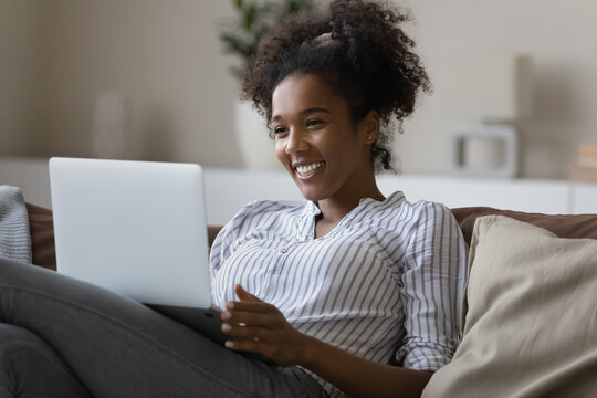 Smiling African American Woman Relax On Sofa Talk Speak On Video Call On Laptop. Happy Young Ethnic Female Have Fun Laugh Engaged In Webcam Digital Virtual Event On Computer. Technology Concept.