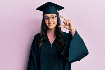 Young hispanic woman wearing graduation cap and ceremony robe smiling and confident gesturing with hand doing small size sign with fingers looking and the camera. measure concept.