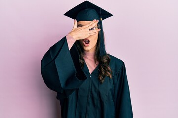 Young hispanic woman wearing graduation cap and ceremony robe peeking in shock covering face and eyes with hand, looking through fingers with embarrassed expression.