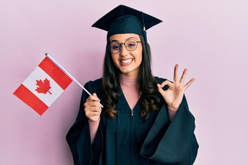 Young hispanic woman wearing graduation uniform holding canada flag doing ok sign with fingers, smiling friendly gesturing excellent symbol