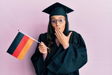 Young hispanic woman wearing graduation uniform holding germany flag covering mouth with hand, shocked and afraid for mistake. surprised expression