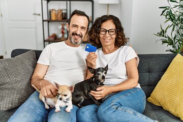 Middle age hispanic couple smiling happy and holding credit card. Sitting on the sofa with dogs at home.