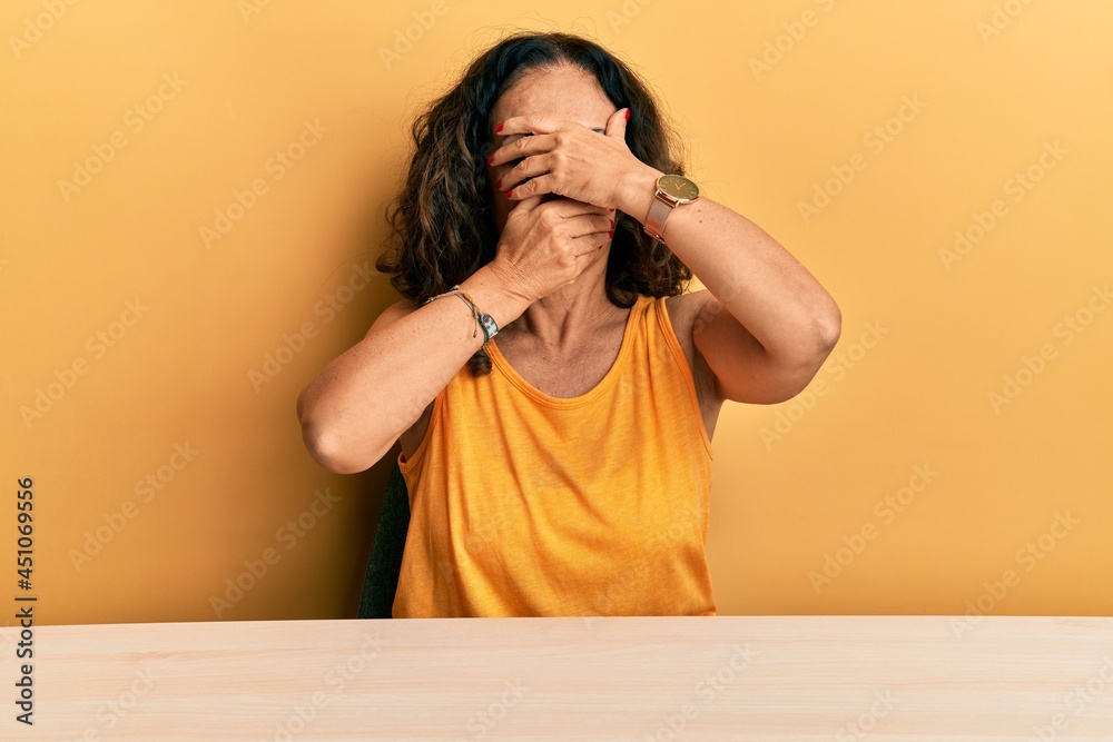 Poster beautiful middle age woman wearing casual clothes sitting on the table covering eyes and mouth with 