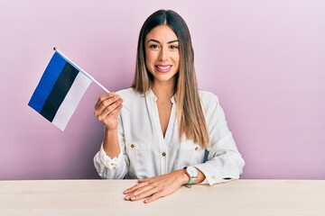 Young hispanic woman holding estonia flag sitting on the table looking positive and happy standing...