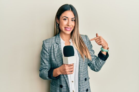 Young hispanic woman holding reporter microphone pointing finger to one self smiling happy and proud