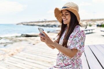 Young latin girl wearing summer using smartphone sitting on the bench hat at the beach.