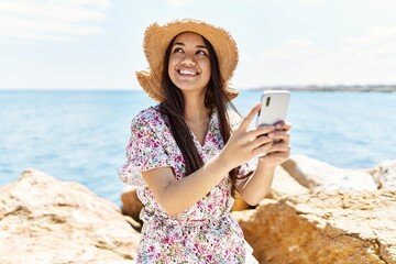 Fototapeta premium Young latin girl wearing summer hat using smartphone at the beach.