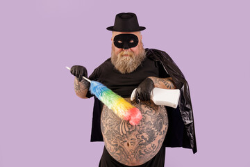 Mature man with overweight in hero costime with large bare tattooed belly sprays detergent onto...