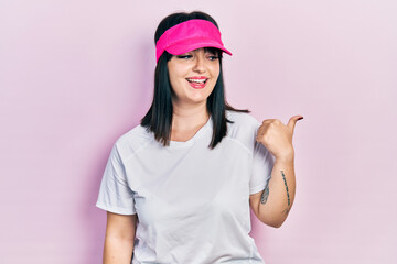 Fototapeta na wymiar Young hispanic woman wearing sportswear and sun visor cap smiling with happy face looking and pointing to the side with thumb up.