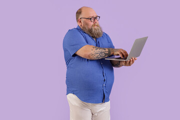 Emotional mature bearded man with overweight in tight shirt with glasses works on laptop standing...