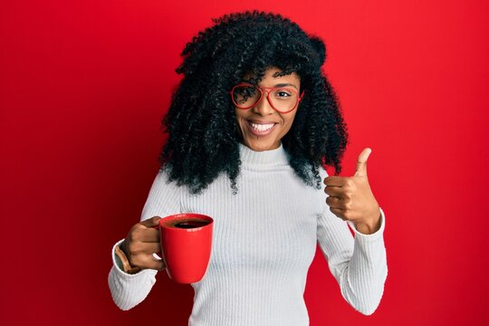 African american woman with afro hair holding coffee smiling happy and positive, thumb up doing excellent and approval sign