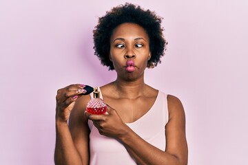 Young african american woman holding perfume making fish face with mouth and squinting eyes, crazy...