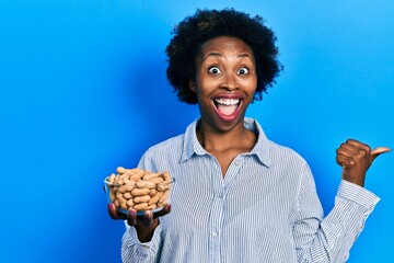 Young african american woman holding peanuts pointing thumb up to the side smiling happy with open mouth